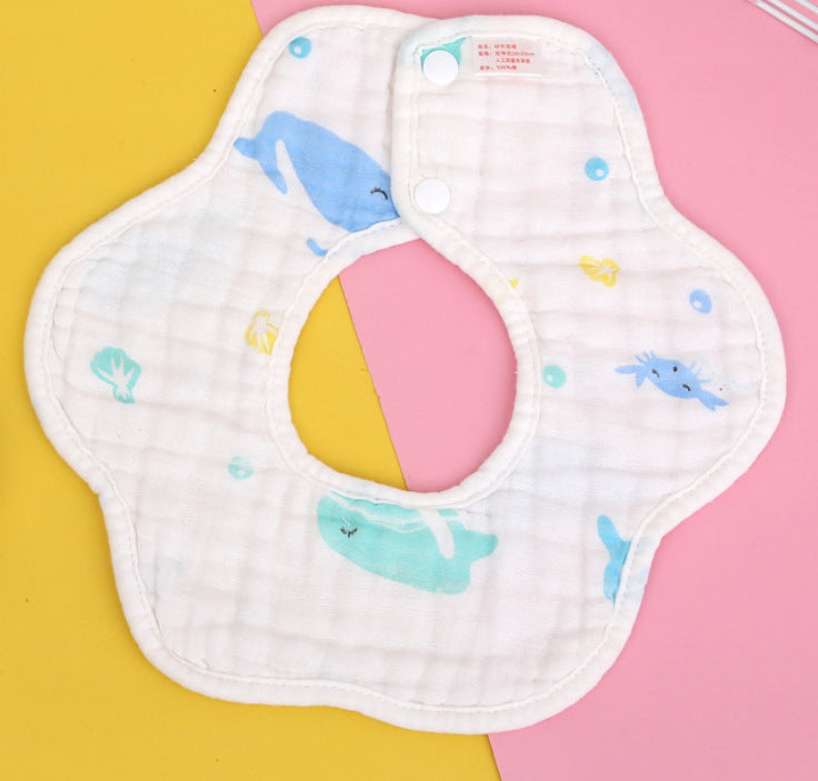 White baby bib with a floral shape, featuring blue and green whale illustrations. The background is pink and yellow, highlighting the bib's playful design.