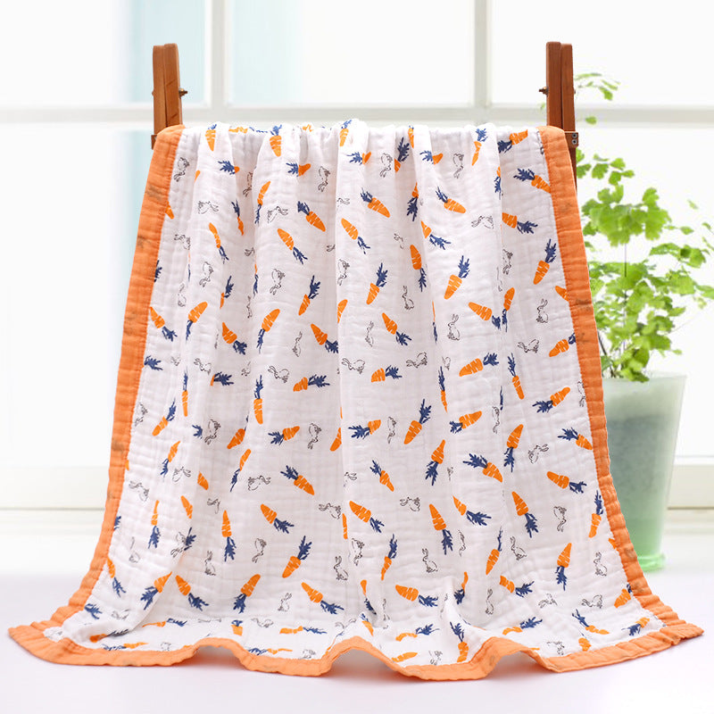 A soft white blanket with orange and blue carrot and bunny patterns, edged in orange, hangs on a wooden rack. A green plant in the background adds a fresh touch.