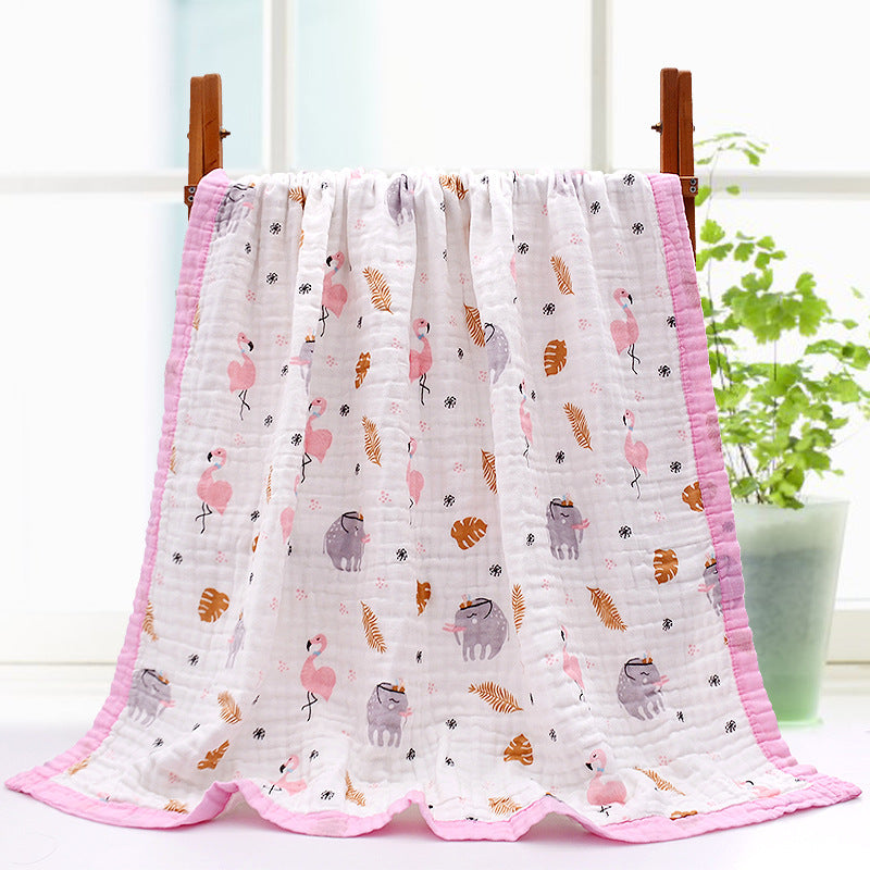 A soft white blanket with pink edges, featuring a pattern of pink flamingos and cartoon sloths, hangs on a wooden rack. A potted plant is visible in the background.