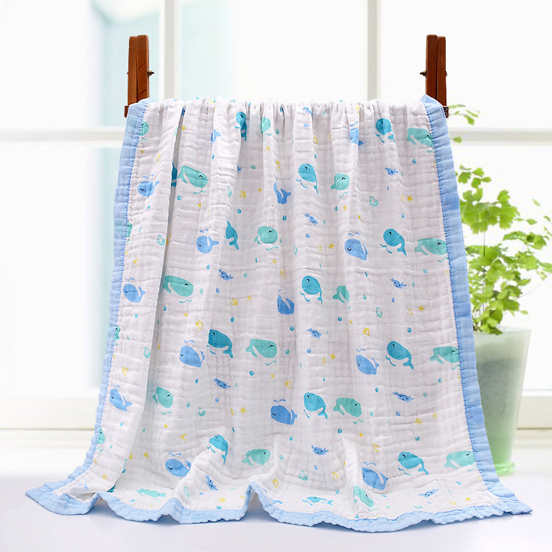 White baby blanket with blue trim featuring cute blue and green cartoon whales and fish. It's draped on a hanger, with a plant in the background.