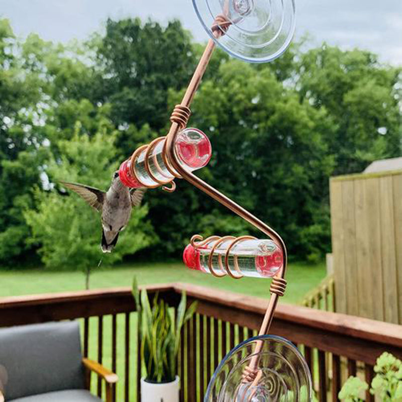 A hummingbird hovers near a red and clear spiral feeder attached to a window with suction cups. A wooden deck with seating and planters is in the background, surrounded by lush trees.