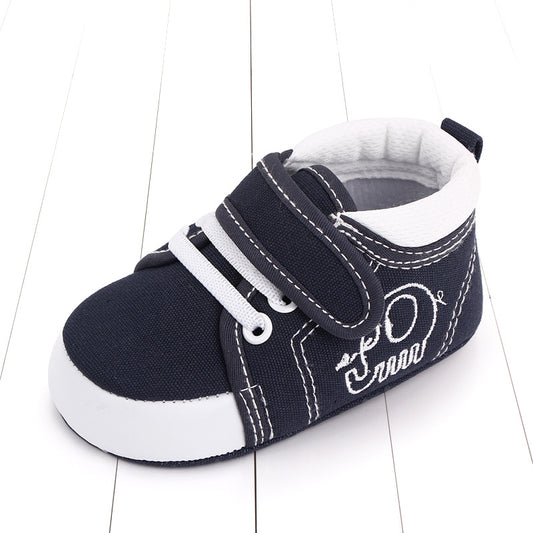 A navy blue baby shoe with white accents and laces, featuring a Velcro strap and embroidered design, placed on a white wood surface.