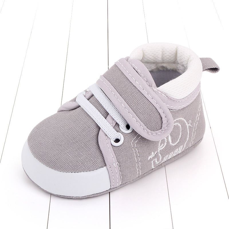 Gray baby sneaker with soft white toe cap, Velcro strap, and laces, resting on a white wooden floor. Embroidered white stitching adds detail to the sides.