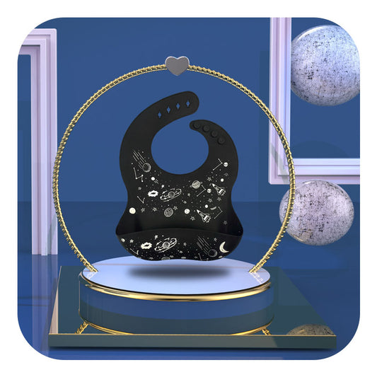 Black baby bib with space-themed illustrations, suspended on a stand with a heart at the top. Background features blue walls, framed sections, and silver spheres.