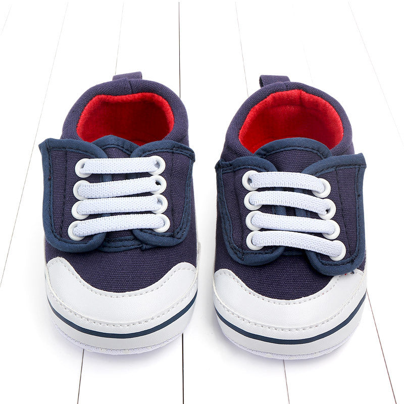 Navy blue baby sneakers with white laces and rubber soles, featuring red interiors, are placed side by side on a white wooden surface.