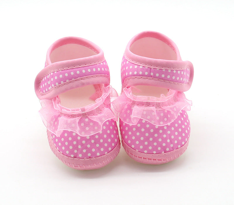 Pink baby shoes with white polka dots, featuring velcro straps and a frilly lace trim around the ankle, displayed on a plain white background.