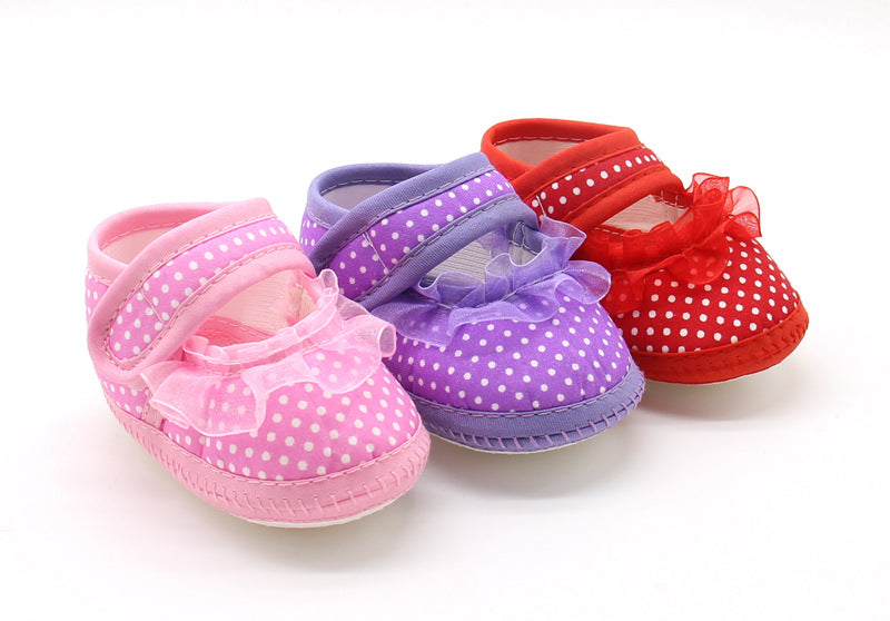 Three baby shoes in pink, purple, and red with white polka dots and sheer ruffle accents are arranged side by side on a white background.