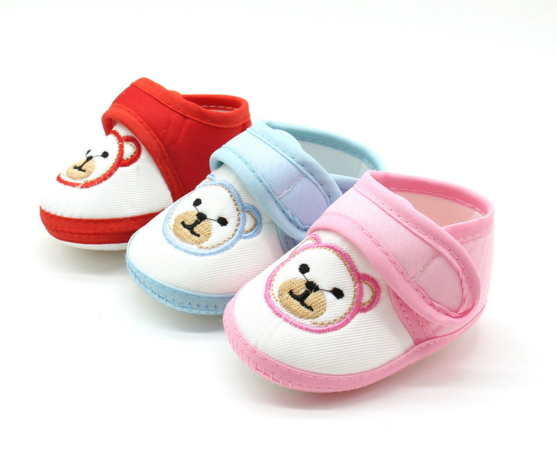 Three pairs of baby shoes in red, blue, and pink, each featuring an embroidered smiling bear face. The shoes have soft, Velcro straps and contrasting colored soles.