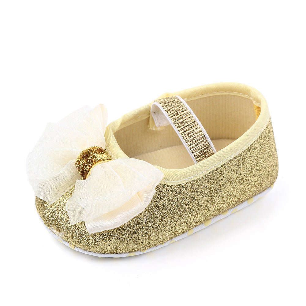 Golden glitter baby shoe with a large white bow, gold center, and a matching glittery strap. The shoe has a soft, cream lining, designed for infants, with an elegant and stylish look.