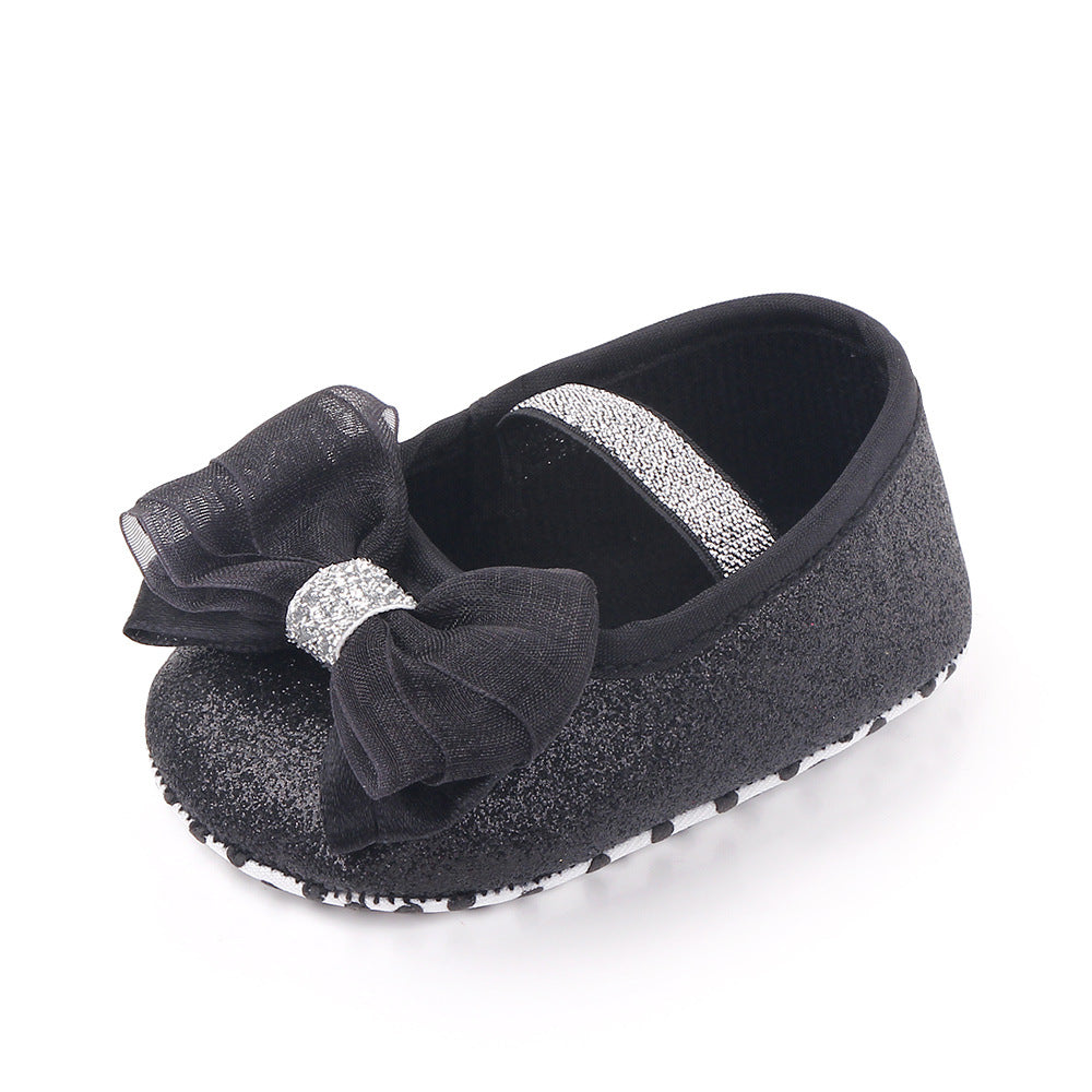 Black baby shoe with sparkly finish, adorned with a large sheer bow and silver glitter strap. The shoe has a soft sole with dotted grips for traction.