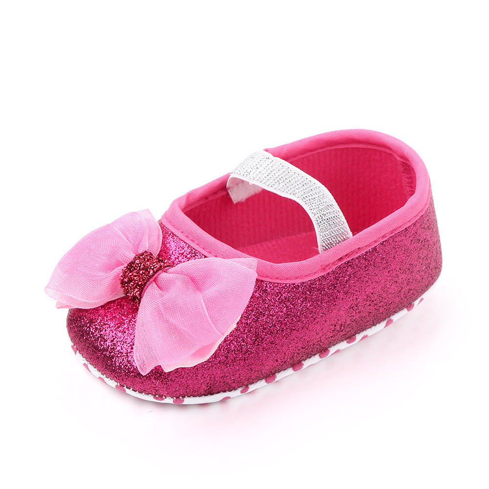 Pink glittery baby shoe with a large bow, featuring a decorative rhinestone center. The shoe has a white elastic strap and a soft, textured white sole for grip.