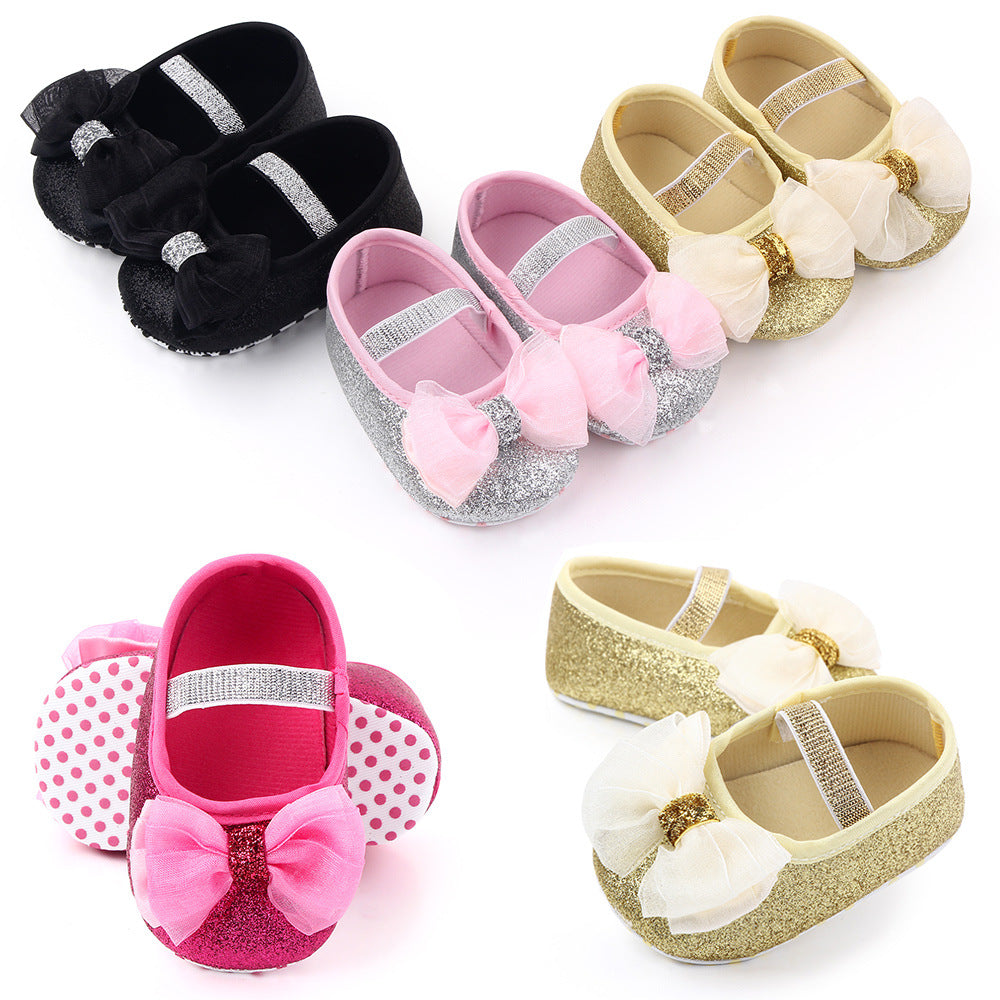 Colorful baby shoes arranged in pairs: black with bow, silver with pink bow, gold with cream bow, and pink with polka dot ears and pink bow.