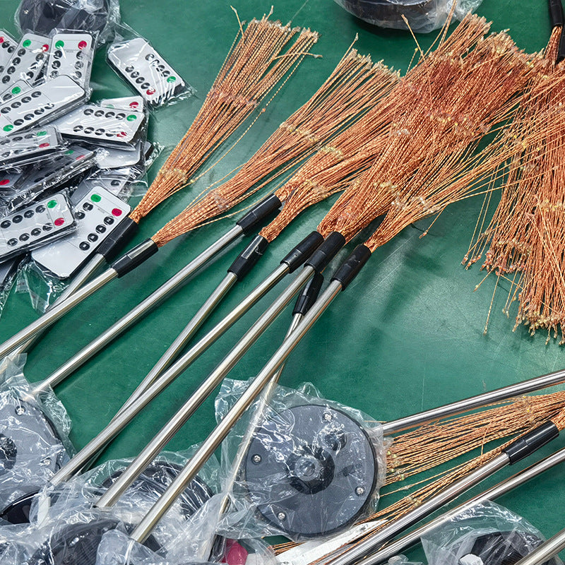 Assembled copper brooms with long handles are laid out on a green surface, next to a stack of packaged remote controls with buttons, including red, green, and black.