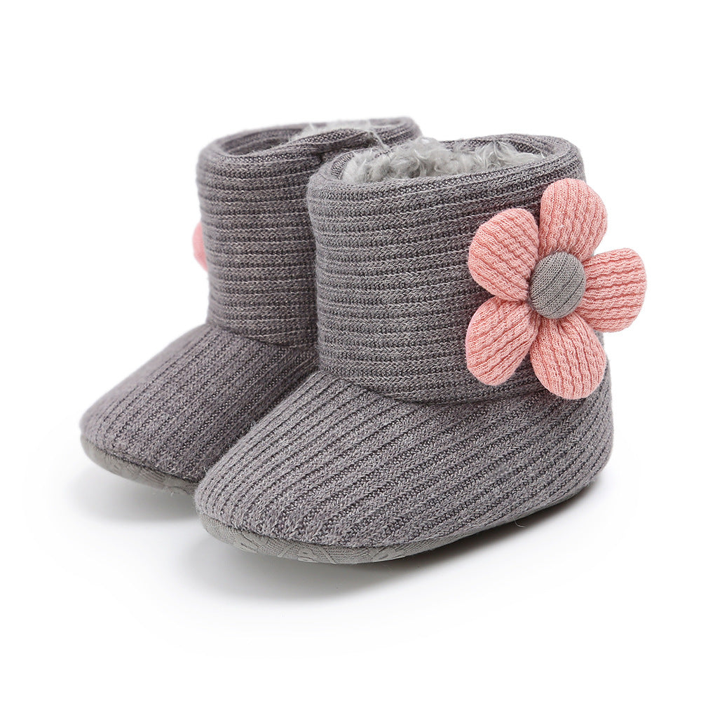 Gray baby boots with ribbed texture and soft lining, each adorned with a large pink fabric flower on the side. The sole is soft and slightly raised at the front.
