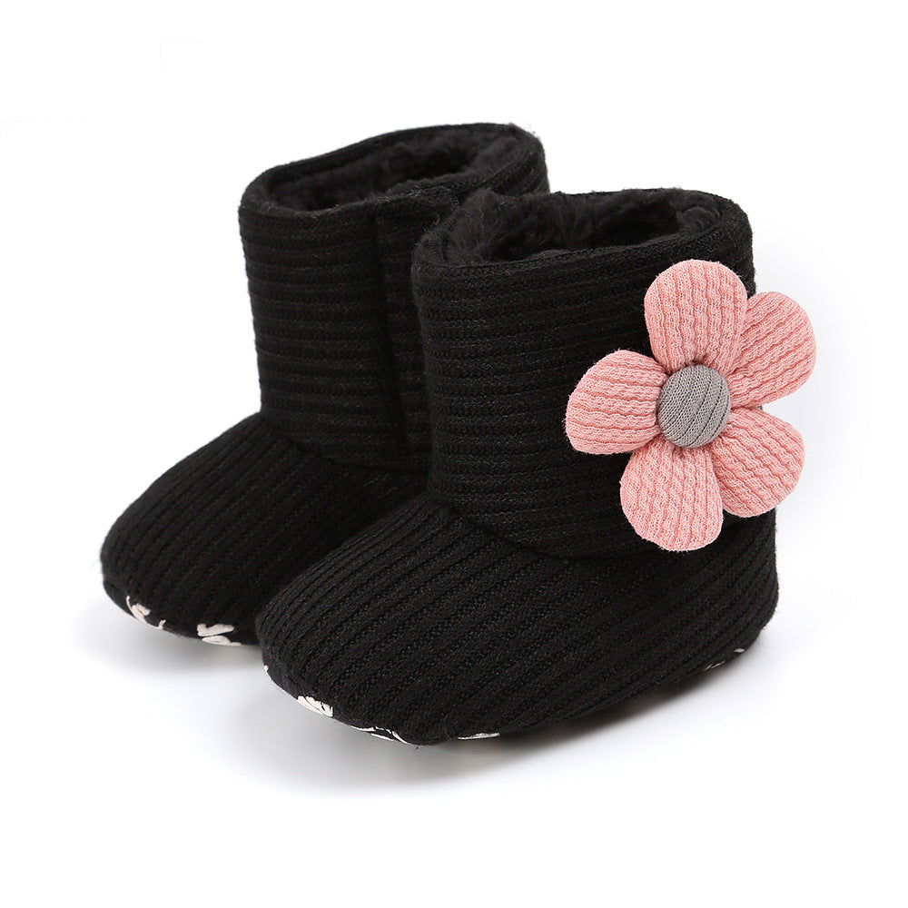 Black knitted baby boots featuring a large pink fabric flower on the side. The boots have a soft, fuzzy interior and rounded toes, providing warmth and comfort.