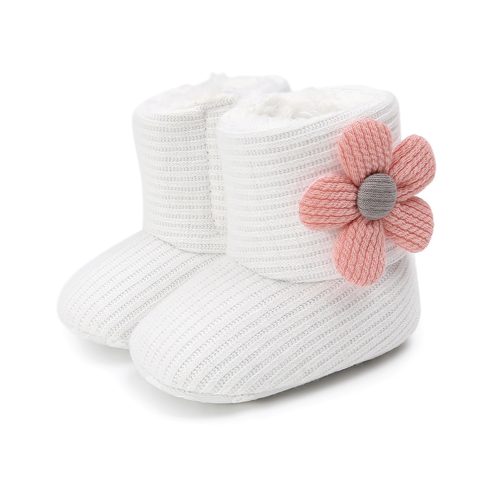 White knitted baby booties with a soft texture, featuring a large pink flower applique on the side. The interior looks plush, providing warmth and comfort.