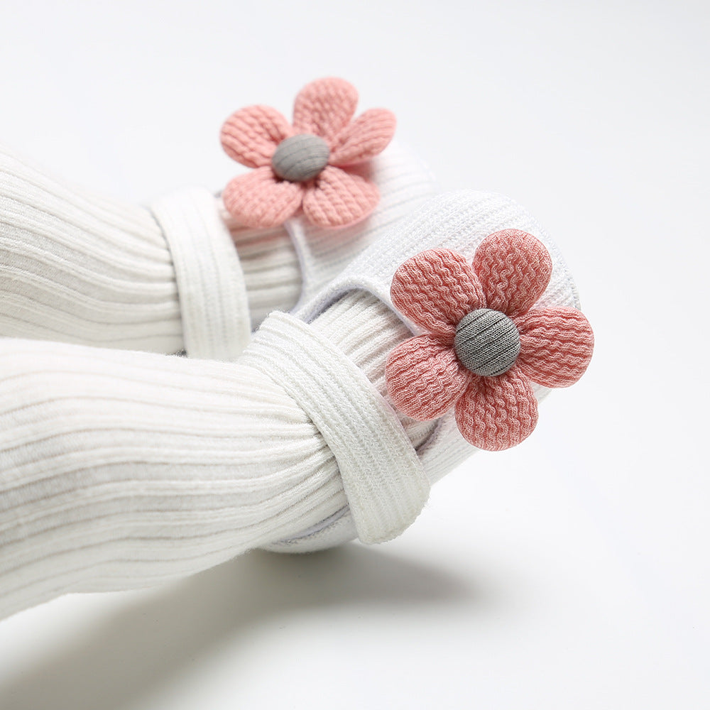 White ribbed fabric shoes with pink knitted flower embellishments featuring gray centers, worn by a child. The shoes are soft with adjustable strap detailing.