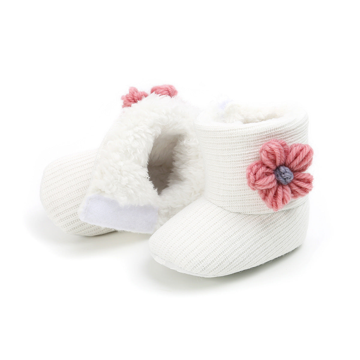 White knitted baby booties with a fluffy interior and pink flower embellishments on the side. One bootie stands upright, while the other shows the soft lining.