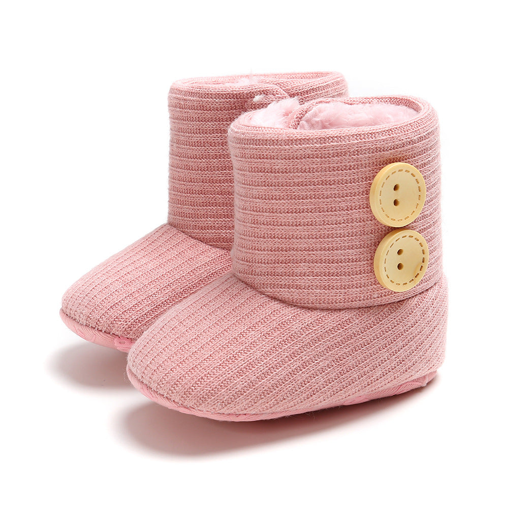 Pink knitted baby booties with ribbed texture, featuring two large yellow buttons on each side. The interior is lined with soft, fluffy fabric for comfort.