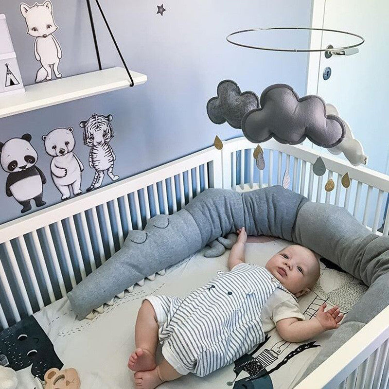 A baby lies in a crib with a blue wall decorated with animal drawings. A soft cloud mobile hangs above. The baby wears striped overalls, surrounded by a gray snake pillow.