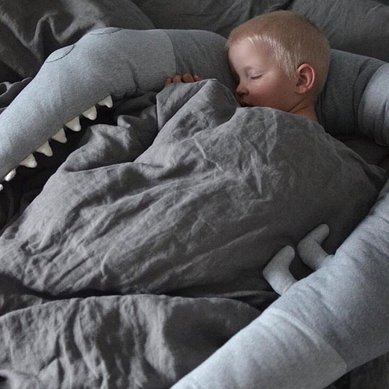 Young child asleep, cuddled in a cozy gray bedding set, which resembles a friendly, plush shark hugging them gently with its plush teeth and fins.