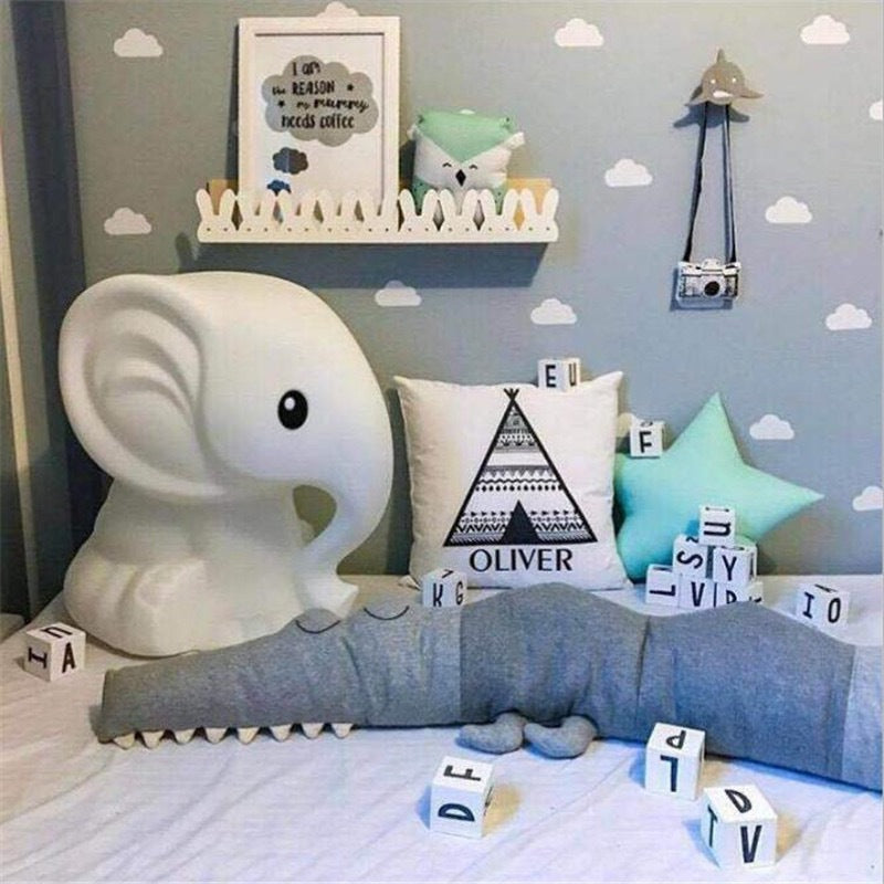 Child's room with a large white elephant lamp, decorative pillows, a plush crocodile, and scattered letter blocks. A shelf holds toys beneath a framed print and cloud wallpaper.