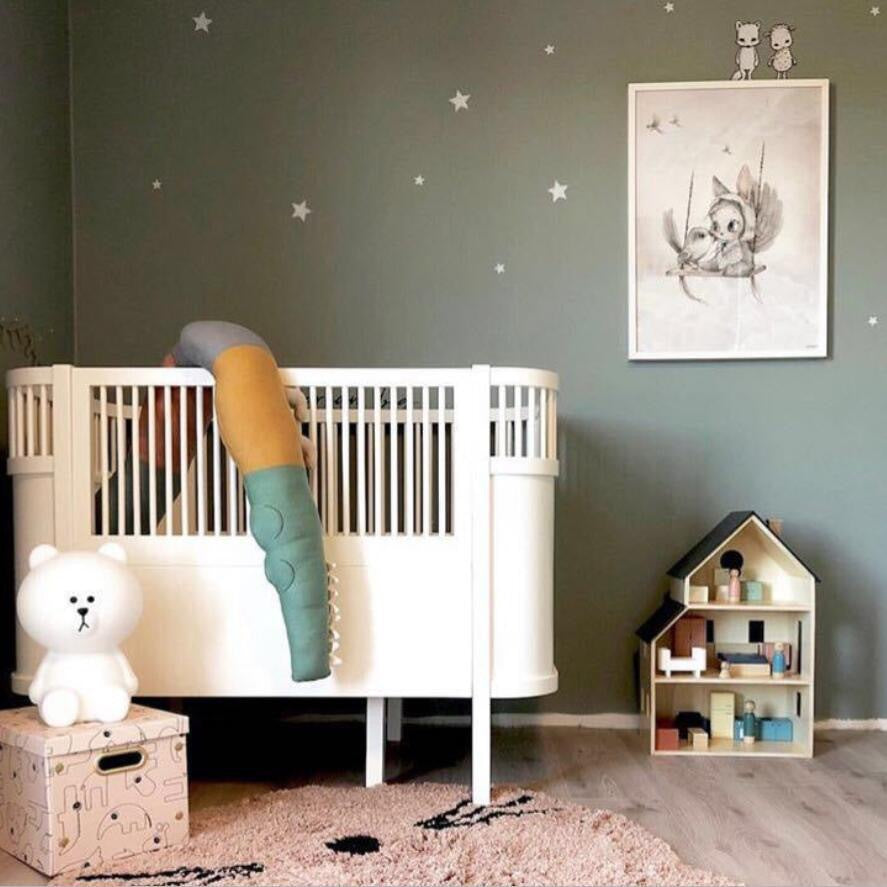 A nursery with a white crib, a colorful stuffed toy, and a bear lamp on a box. The walls are green with star decals, featuring a framed animal drawing and a dollhouse shelf.