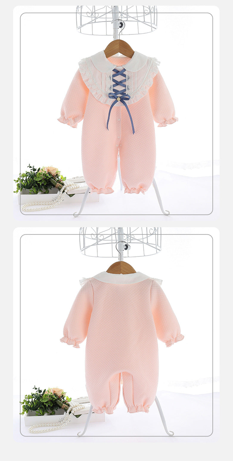 Pink baby romper with lace collar and a blue ribbon detail at the front, displayed on a hanger. Features ruffled cuffs and a soft quilted pattern, with floral decor nearby.