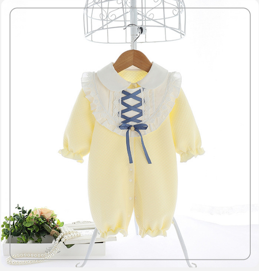 A yellow baby romper with a white, ruffled collar and blue ribbon lacing on the front. It has long sleeves with ruffled cuffs and is displayed on a stand with decorative items nearby.