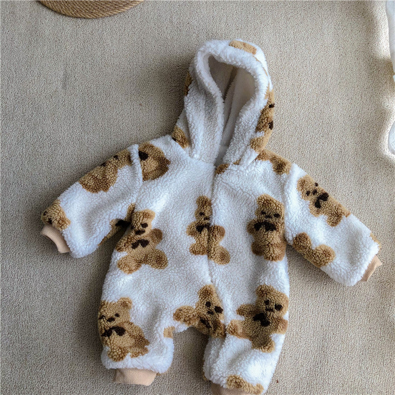 Fluffy white baby jumpsuit with teddy bear print on light brown fabric, featuring a hood and long sleeves, lying flat on a textured beige surface.