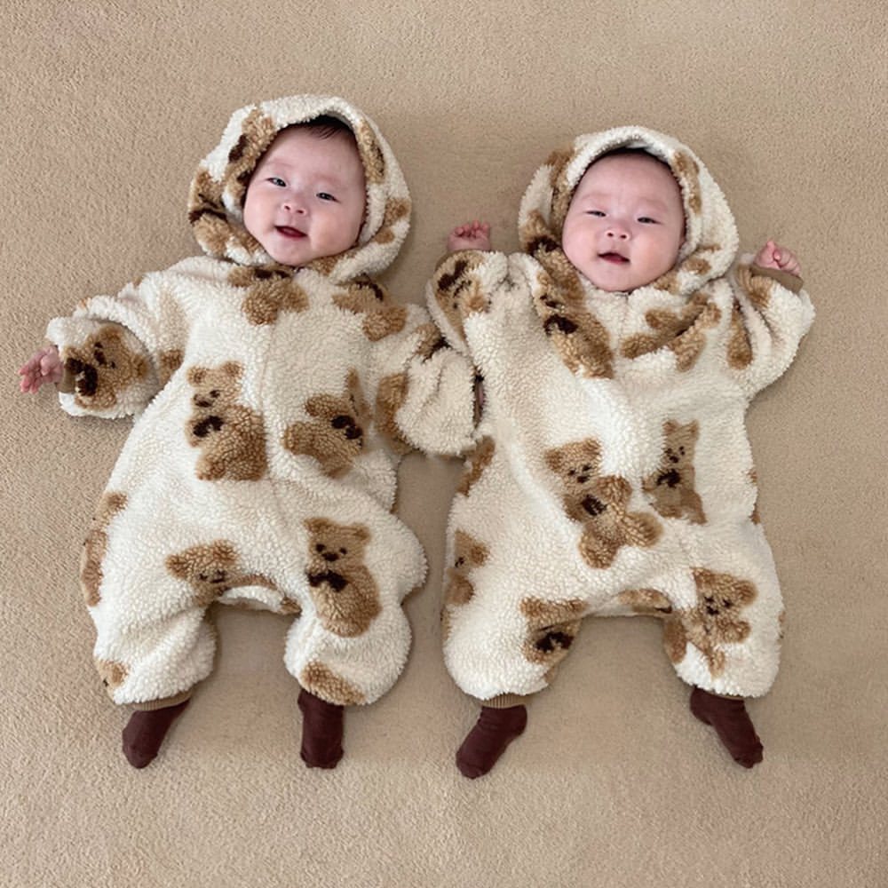 Two adorable babies lying on a beige surface, wearing matching fluffy onesies with teddy bear patterns and hoods. They have small, happy expressions and cozy brown socks.