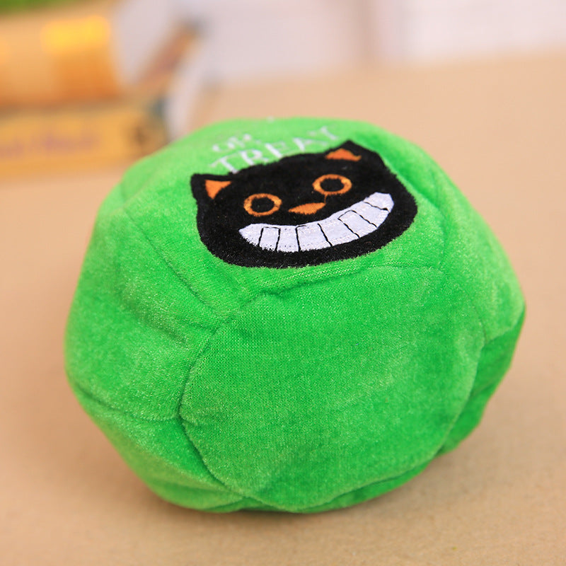 A green plush ball with a black smiling cat face design, featuring orange eyes and pointy ears. The background is blurred, showcasing books and a light surface.
