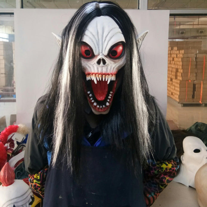 Person wearing a scary mask with long black hair, pointy ears, red eyes, and sharp teeth, standing indoors with other masks and materials visible in the background.