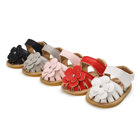 Five pairs of baby sandals in black, grey, pink, red, and white. Each pair features a decorative flower on the front, with a soft sole and an open toe design.