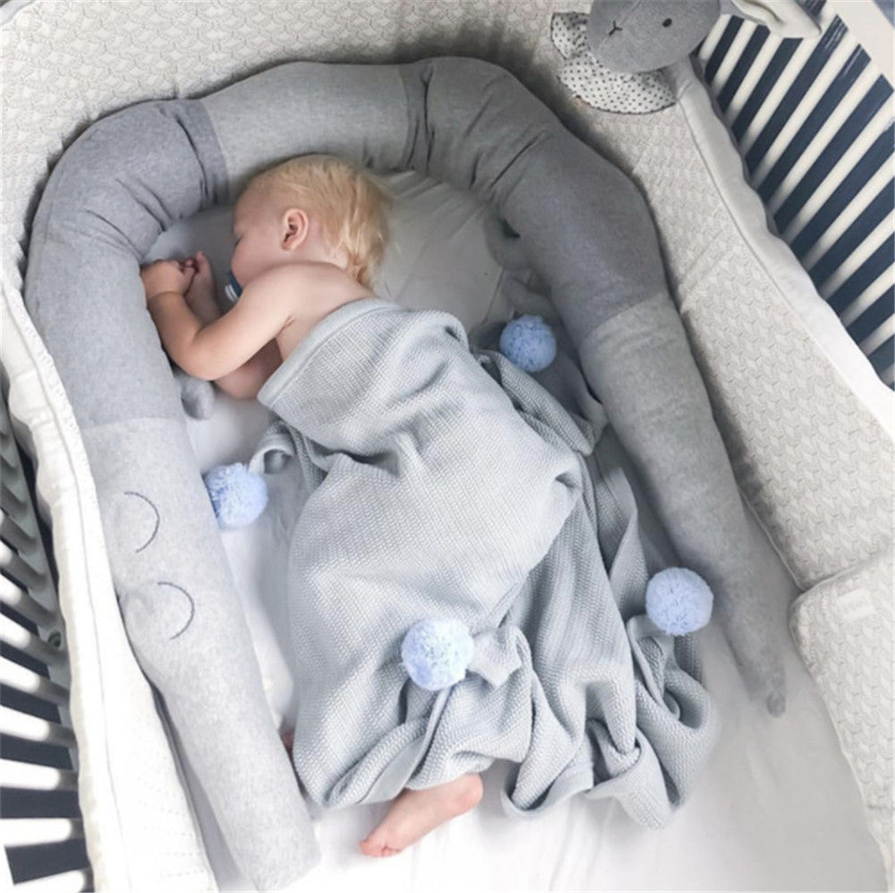 A baby sleeps in a crib, surrounded by a gray snake-shaped cushion. The baby is covered with a light gray blanket adorned with blue pom-poms, next to a plush rabbit toy.