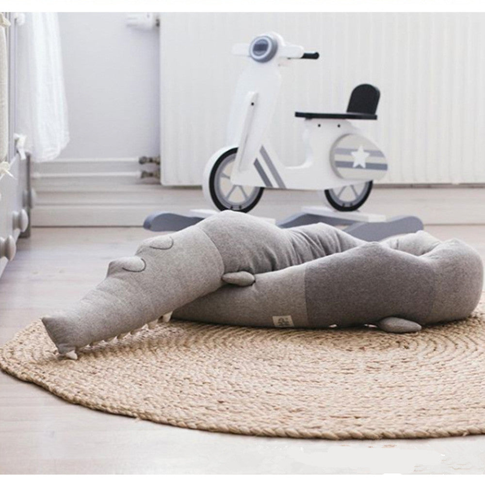 Soft, gray crocodile-shaped pillow on a woven rug in a minimalist nursery, with a white wooden toy scooter in the background, near a radiator.