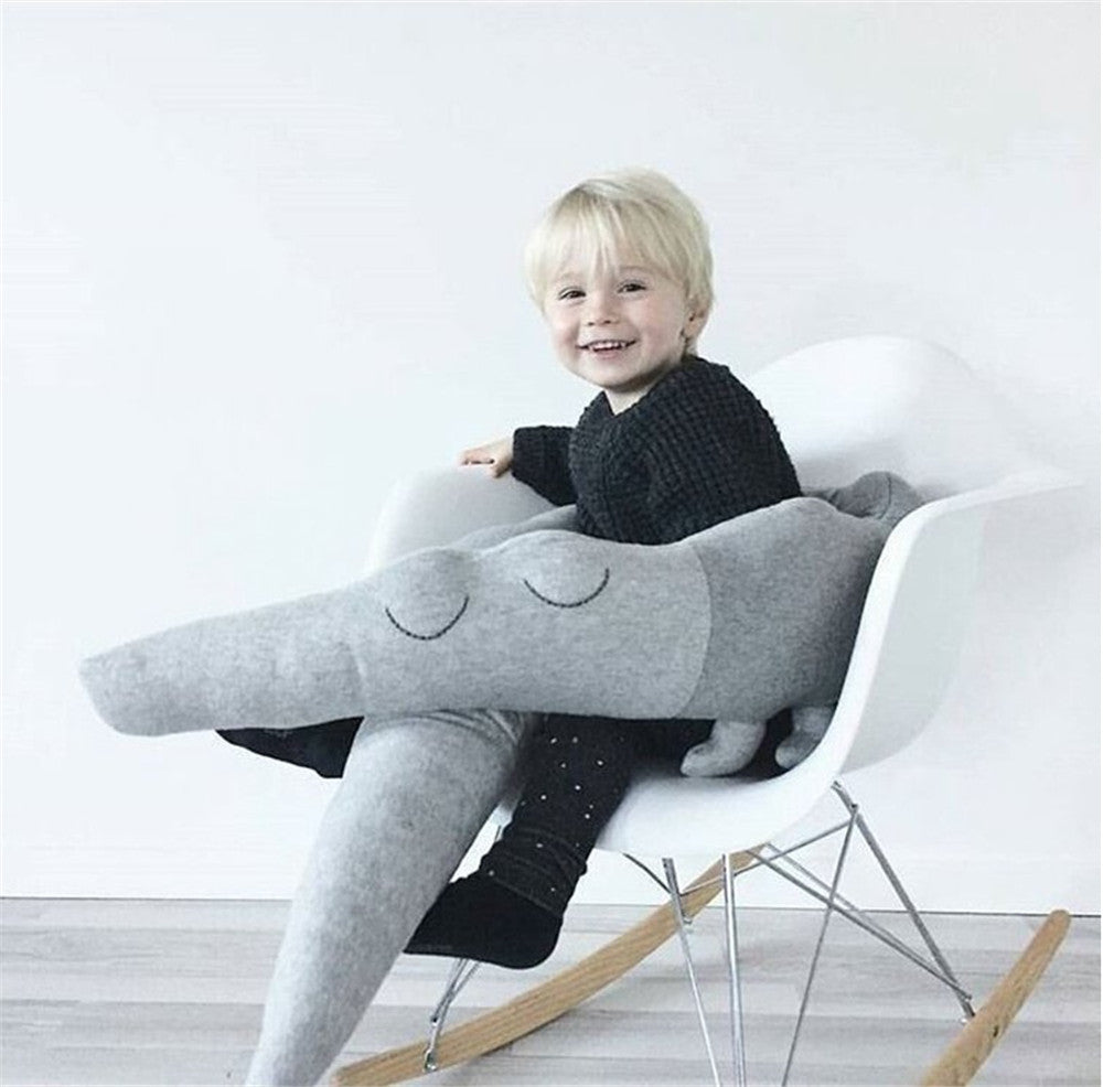 A happy child sits on a white rocking chair, hugging a large, plush gray dinosaur toy. The room has a minimalist design with light wood flooring and a plain white wall.
