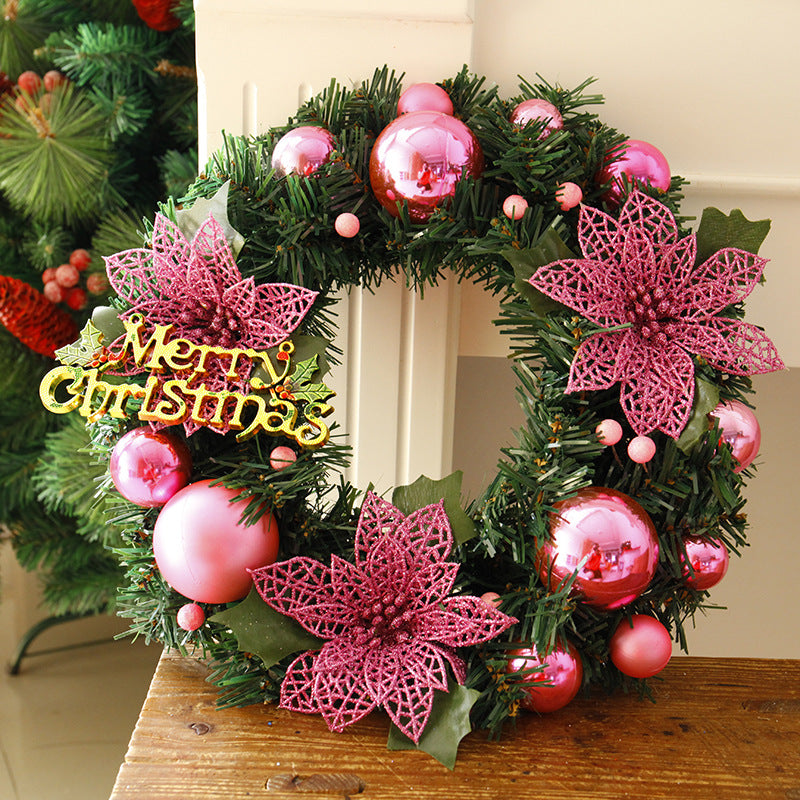 Christmas wreath with pink poinsettias, shiny pink baubles, and a "Merry Christmas" sign, displayed on a wooden table near a decorated tree.