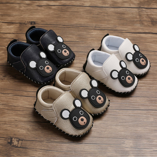 Three pairs of baby shoes on a wooden surface, each featuring a cute bear face. The shoes are black, beige, and white, with contrasting soft soles and rounded ears.