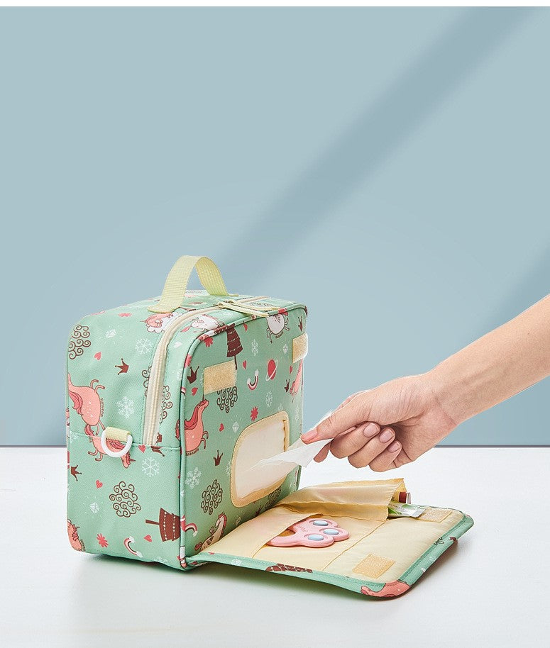 A hand reaches into a pastel green diaper bag adorned with playful animal patterns and snowflakes. The front flap is open, revealing a tissue dispensing pocket and diaper storage.