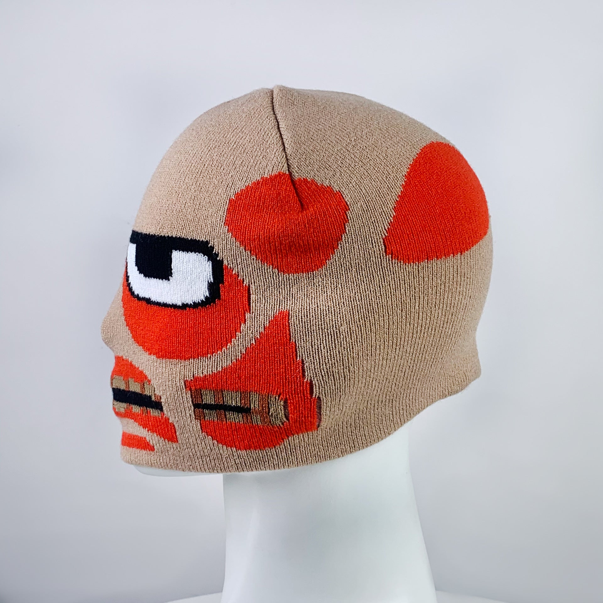 Beige knit beanie with abstract red and black face design, featuring a large eye and geometric shapes. Displayed on a white mannequin head against a plain backdrop.