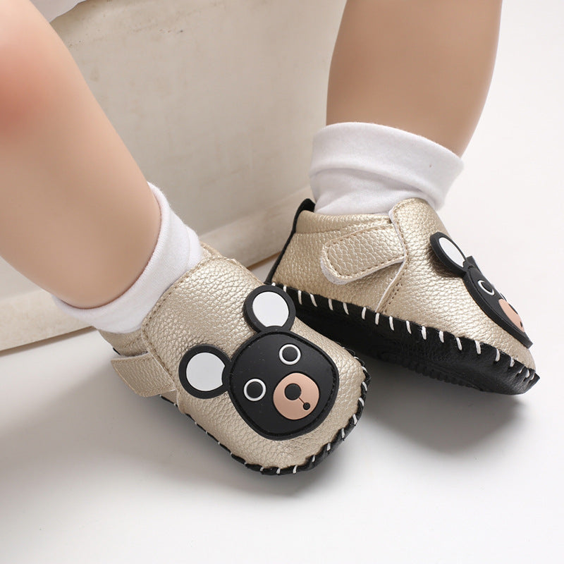 Baby wearing golden soft-soled shoes featuring a cute black bear face with white ears, paired with white socks, sitting on a light surface.