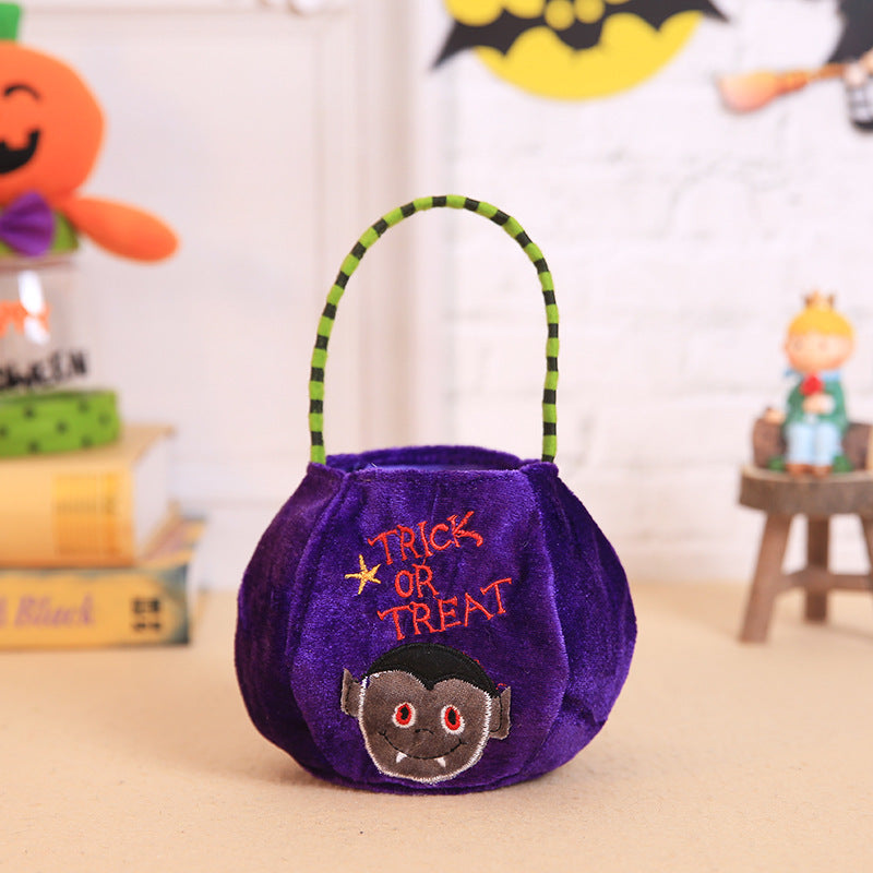 A purple trick-or-treat bucket with a vampire face design and a green striped handle sits on a table. Halloween decorations, including a pumpkin, are blurred in the background.
