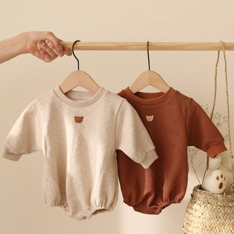 Two baby rompers hang on a wooden rod. One is beige, and the other is brown, both adorned with a small bear face patch. A hand holds the rod, with a woven basket in the background.