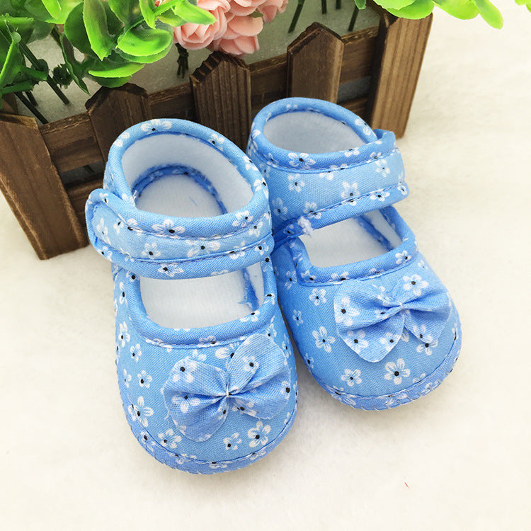 Blue baby shoes with white floral patterns and decorative bows on each toe, placed on a white surface. A small wooden fence and pink flowers are in the background.