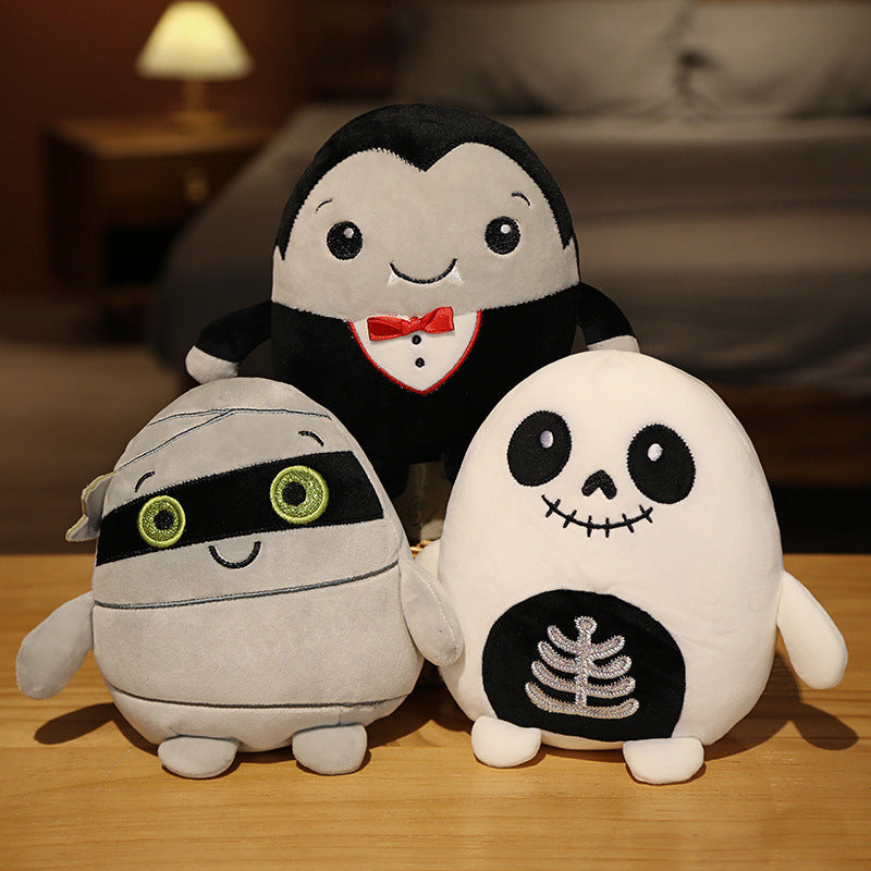 Three plush toys on a wooden surface: a mummy with green eyes, a vampire with a red bow tie, and a skeleton with a black mouth and ribcage detail. Background is a cozy bedroom.