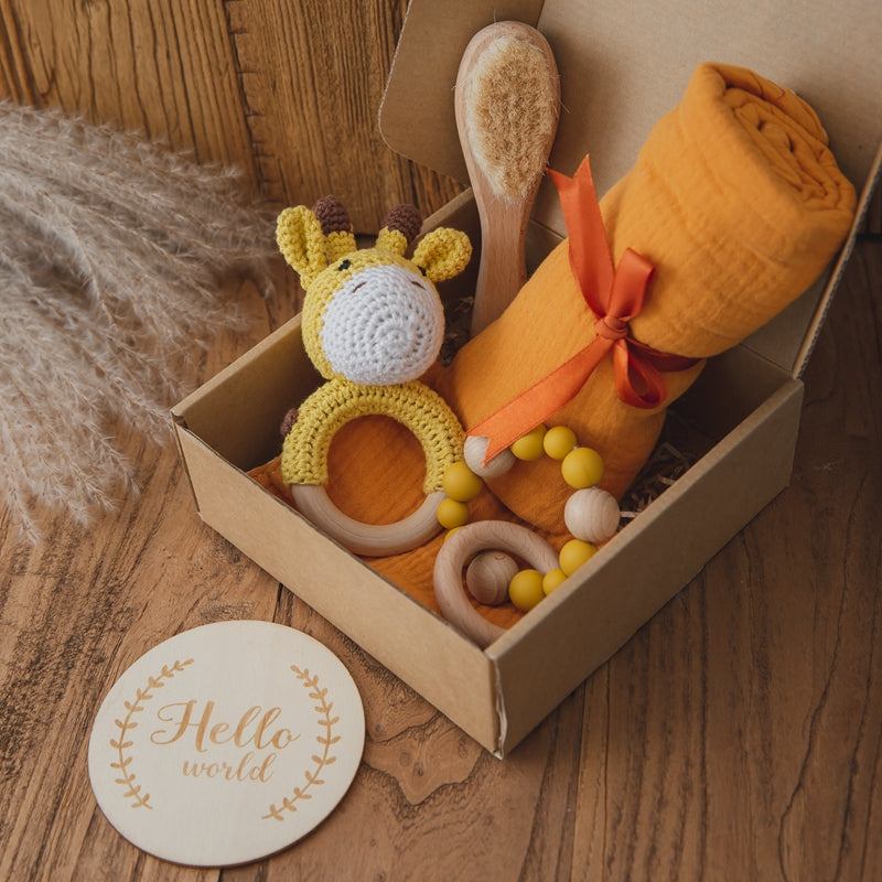 Gift box with an orange blanket, plush giraffe toy, wooden baby brush, and yellow beaded teether. Nearby, a round wooden plaque reads “Hello world” on a wooden surface.