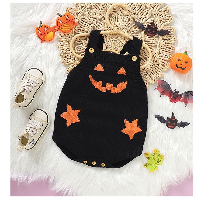 A black romper with orange jack-o'-lantern and star designs is placed on a woven mat. Nearby are Halloween-themed decorations, sunglasses, and a pair of white sneakers.
