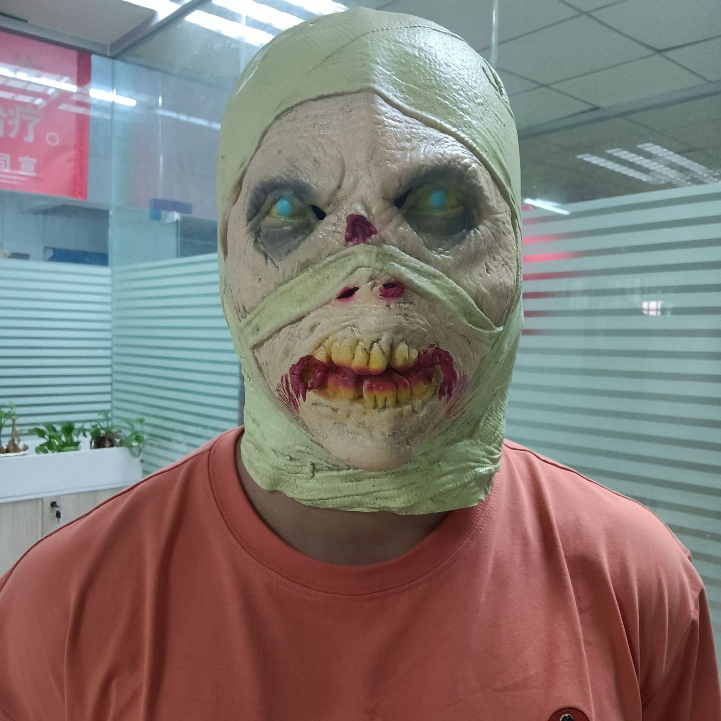 Person wearing a ghoulish, bandage-like mask with decayed features, stands in an office wearing a salmon-colored shirt. Background shows glass walls and a ceiling with lights.