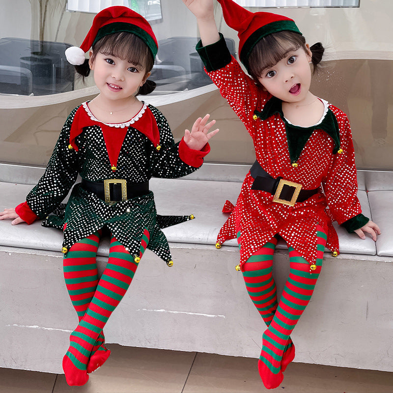 Two children dressed as elves sit on a bench. One wears a green outfit, the other red. Both have matching striped tights and festive hats, smiling and playful.